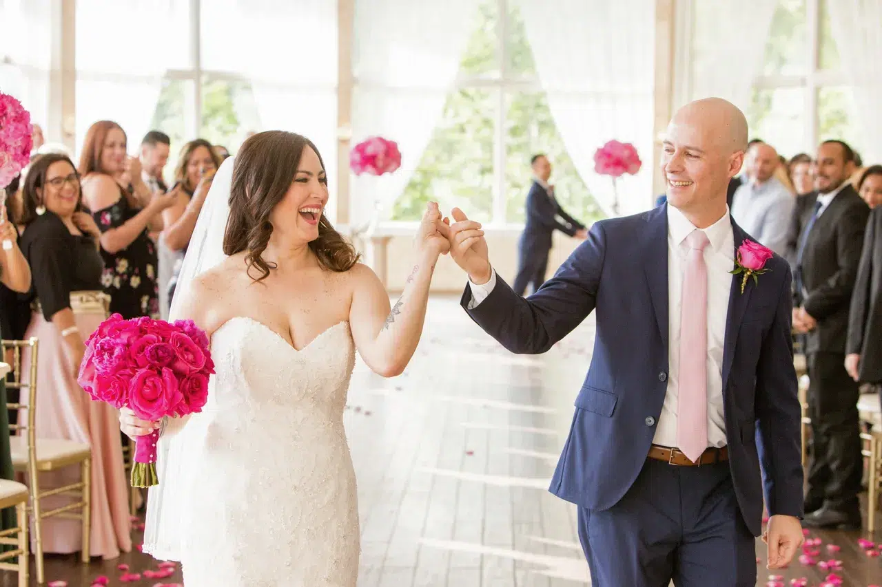 Bride and groom celebrate their marriage, holding hands and smiling, with the bride holding a bouquet of vibrant pink peonies.
