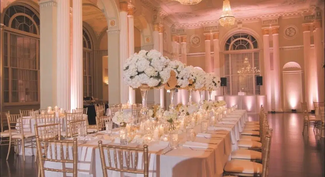Elegant ballroom with long banquet tables, gold chairs, and tall white floral centerpieces.