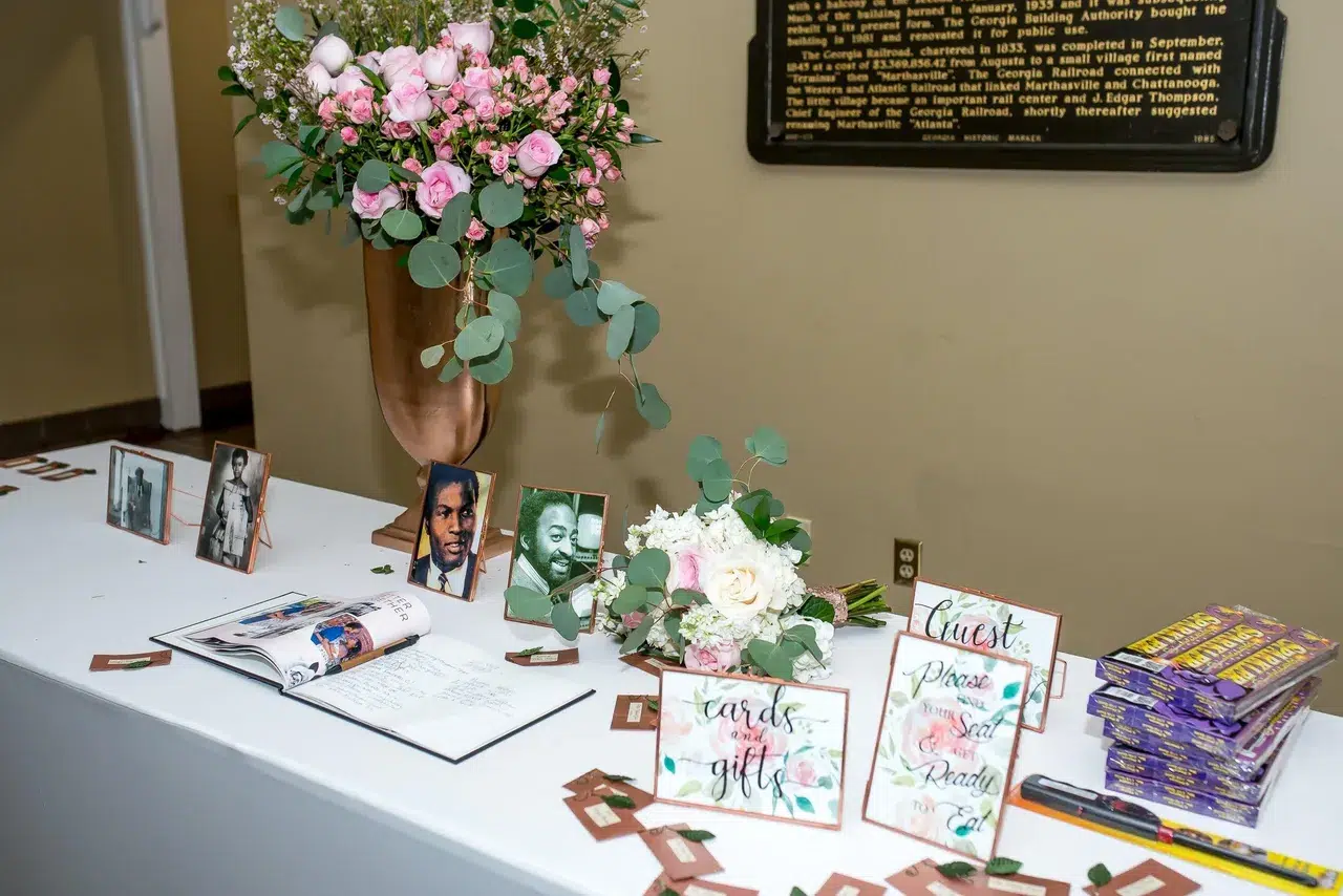 Decorated table at an event featuring floral arrangements with pink roses, photographs of influential figures, and guest amenities like a guest book and cards
