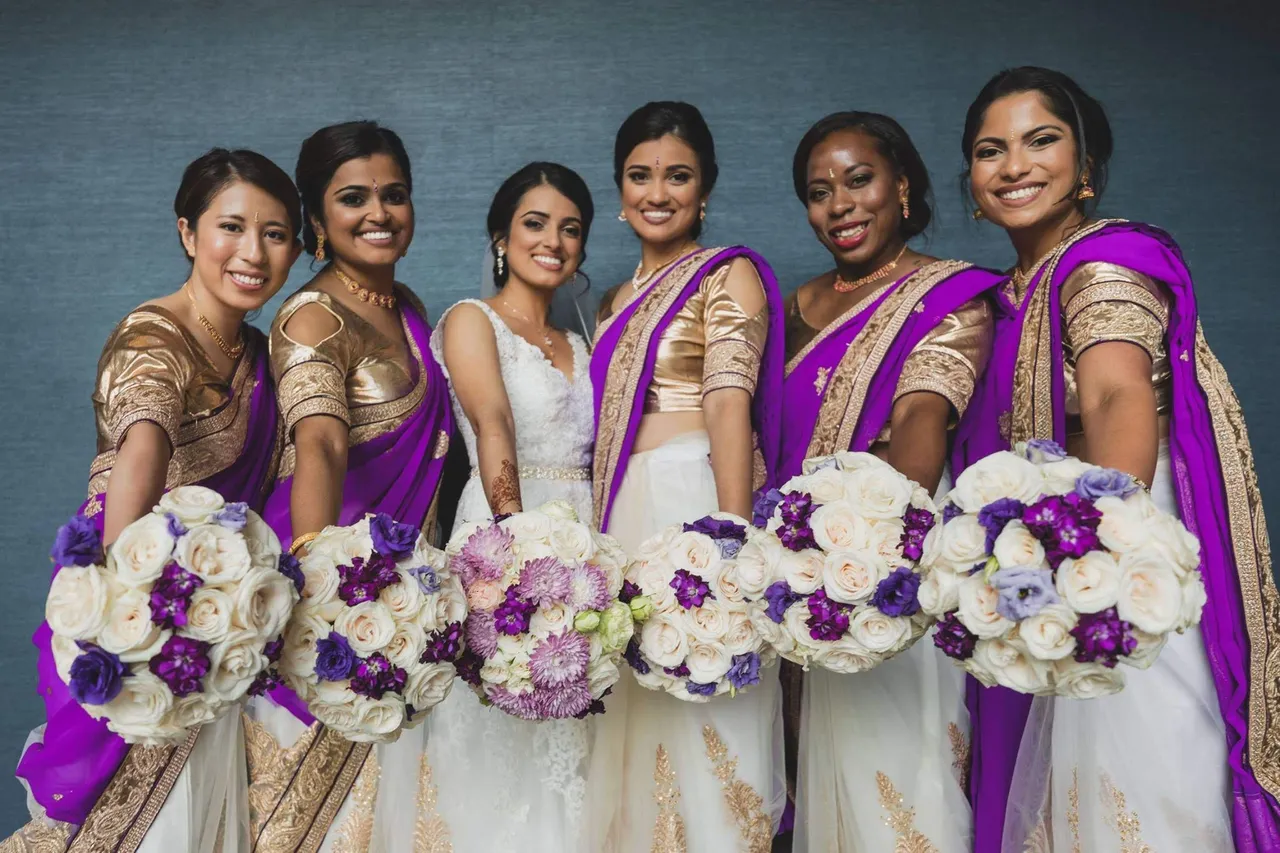 A bride and her bridesmaids at an Indian wedding, all dressed in traditional attire and holding beautifully coordinated bouquets with white and purple flowers.