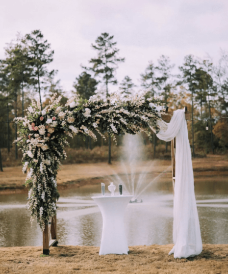 A beautifully designed wedding arch decorated with Atlanta custom wedding flowers, overlooking a tranquil pond with a fountain, set in a serene outdoor setting.