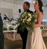 A bride, dressed in a flowing white gown, walks down a rose-petal-lined aisle with her father. Guests are seated on either side, and a draped arch with greenery adorns the end of the aisle at The Stave Room, Atlanta.