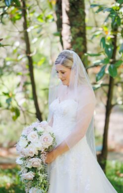 Bride holding a cascading bouquet of blush and ivory roses, standing serenely in a sunlit forest.