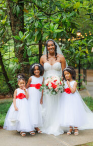 Bride holding a bouquet, accompanied by three young girls in white dresses with red sashes, all smiling.