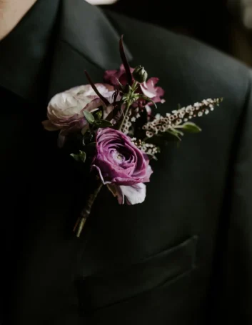 Boutonniere featuring purple wedding flowers pinned to a dark suit jacket, perfect for a sophisticated wedding look.