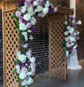 A rustic wooden arch adorned with purple and white Fall wedding flowers, set against an exposed brick backdrop.