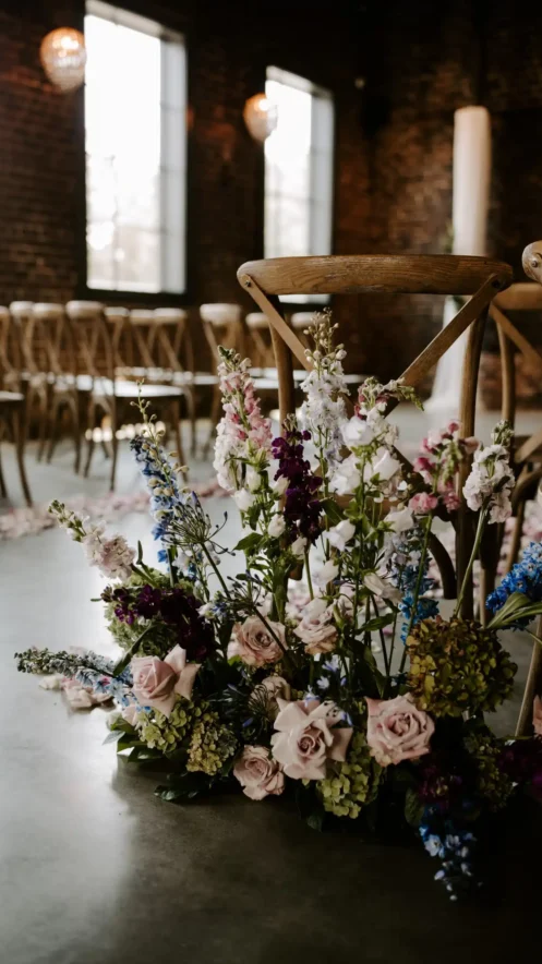 Floral arrangement of pastel roses, hydrangeas, and wildflowers lining the aisle at a Fall wedding ceremony in an elegant indoor venue.