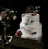 A white wedding cake adorned with blue butterflies and surrounded by Fall wedding flowers on a rustic cake stand.