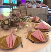 Beautiful wedding table centerpiece with pastel flowers, pink napkins, and gold plates in an elegant reception setting.