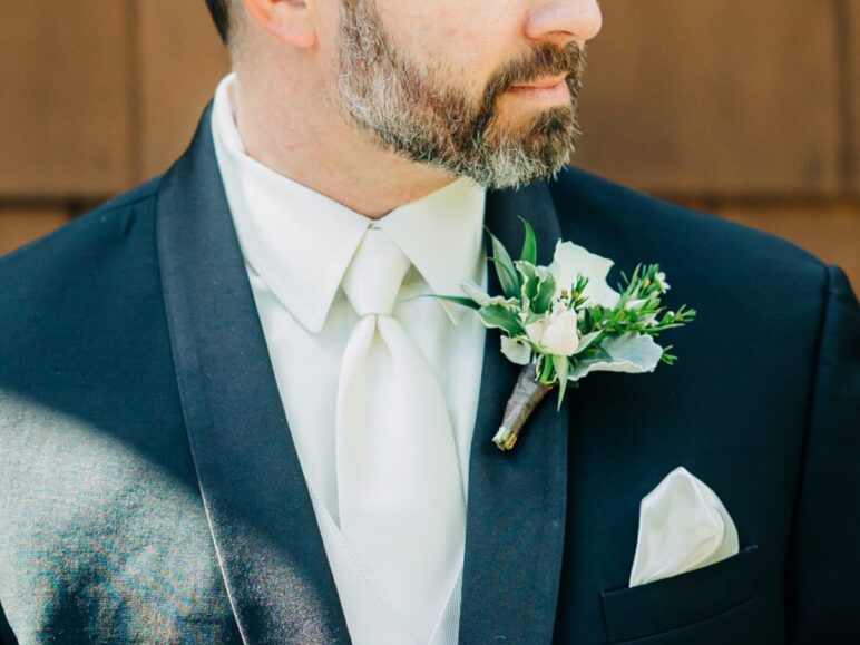 A groom stands against a wooden wall wearing a black suit with a white tie and boutonnieres on his lapel.