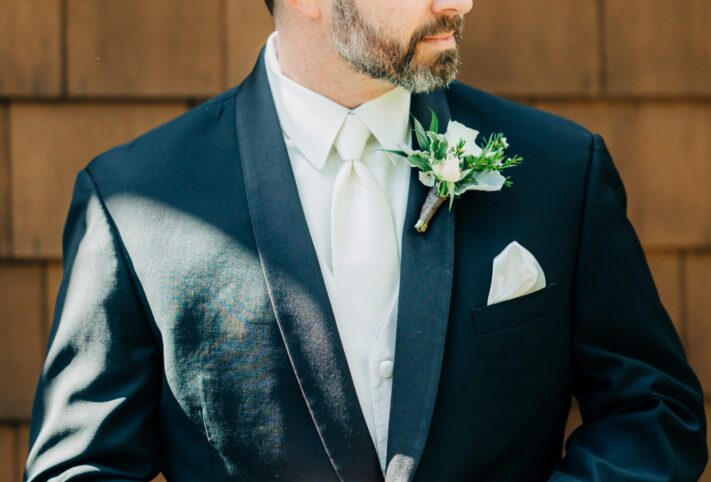 A groom stands against a wooden wall wearing a black suit with a white tie and boutonnieres on his lapel.