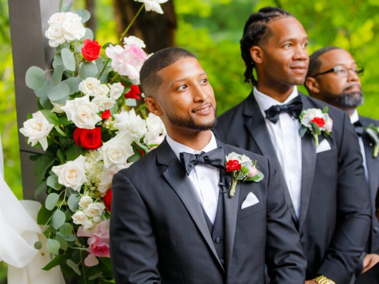 Groomsmen standing attentively during a wedding ceremony, each wearing boutonnieres with white and red flowers