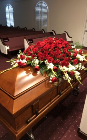 A casket adorned with a large arrangement of red roses and greenery, displayed in a church setting.