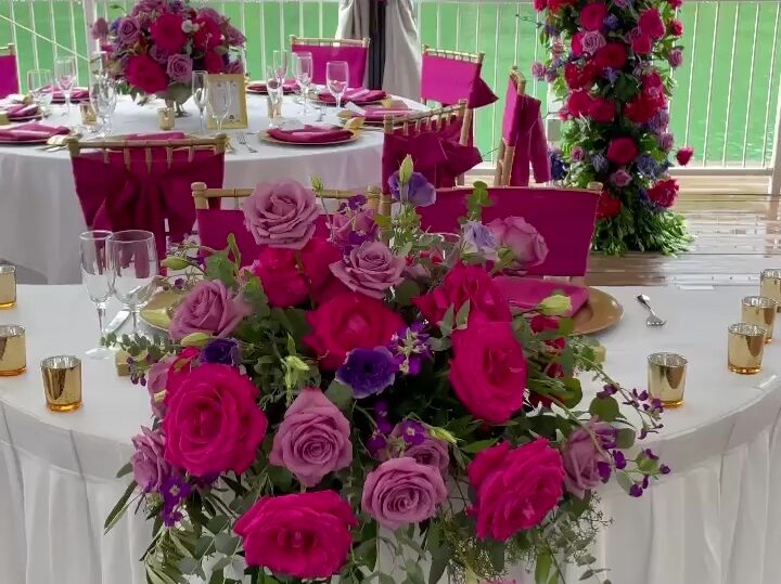 A wedding reception setup featuring a table adorned with vibrant fuchsia pink, hot pink, and purple floral arrangements, with a scenic lake view in the background.