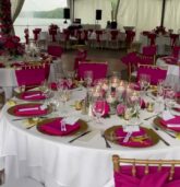 A wedding reception table setup with vibrant fuchsia pink accents, floating candles centerpiece, and elegant table settings under a tent with string lights, overlooking a lake.