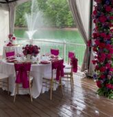 A wedding reception table with fuchsia pink accents, a floral arch, and a lake fountain view, set under a tent