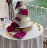 A white tiered wedding cake adorned with vibrant fuchsia pink, purple, and hot pink flowers, set on a table overlooking a lake fountain.