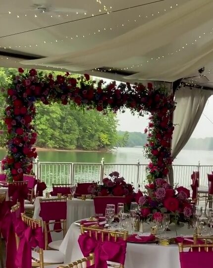 A wedding reception setup featuring tables adorned with vibrant fuchsia pink, hot pink, and purple floral arrangements under a tent with string lights, overlooking a serene lake.