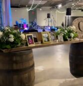 A rustic memory table adorned with white floral arrangements and framed photographs, set on wooden barrels at The Stave Room, Atlanta. String lights and a band stage in the background.