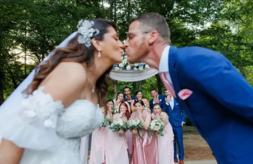 Bride and groom kissing with their bridal party in the background, holding floral bouquets at a summer wedding by Buford Florist.