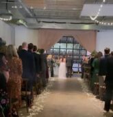 A bride walks down a petal-strewn aisle lined with wooden chairs at The Stave Room, Atlanta. The ceremony backdrop features soft pink drapes, greenery accents, and glowing lanterns.