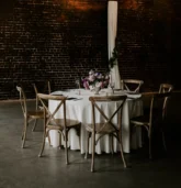 A round reception table with elegant Fall wedding flower decorations, featuring purple and cream blooms, set under warm hanging lights in a rustic venue.