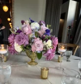 Elegant Fall wedding flower centerpiece featuring roses and seasonal blooms in soft purple and cream, surrounded by candlelight on a reception table.