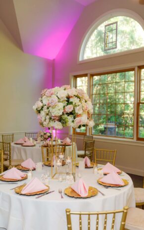 Elegant floral arrangement of white and blush roses and hydrangeas in a tall gold stand, adorning a round reception table set with pink napkins, gold chargers, and votive candles.