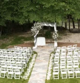 Outdoor wedding ceremony setup with white chairs and a floral arch in a forested area near a river.