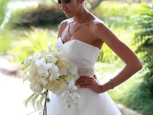 A bride holding a cascading bouquet of white flowers and greenery, standing in a lush, outdoor garden setting.