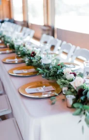 A long wedding table with elegant place settings and a floral and greenery runner decorated with candles