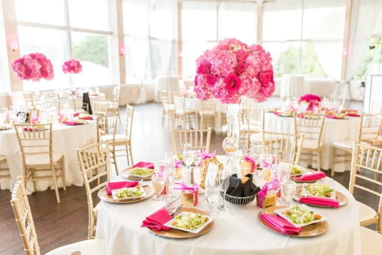 stunning centerpiece of vibrant pink flowers, predominantly roses, arranged in a large, round bouquet atop a tall, clear glass vase.