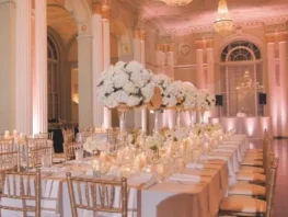 Elegant ballroom with long banquet tables, gold chairs, and tall white floral centerpieces.