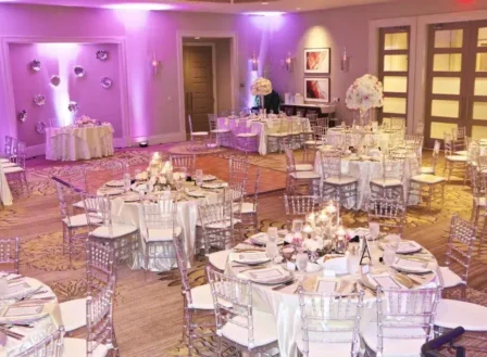 Elegant wedding reception hall with round tables, clear acrylic chairs, and a romantic pink and purple lighting scheme.