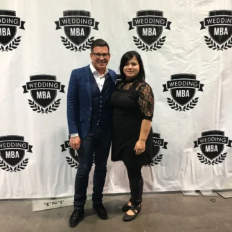 Dina C Tacu and David Tutera posing together at the Wedding MBA event, standing in front of a backdrop featuring the event's logo.