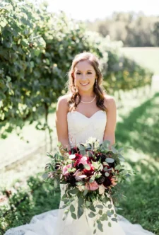 A radiant bride in a vineyard setting, provided by wedding floral services, smiling and holding a large, artfully arranged bouquet of deep red and pink flowers, surrounded by lush greenery.