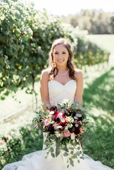 A radiant bride in a vineyard setting, provided by wedding floral services, smiling and holding a large, artfully arranged bouquet of deep red and pink flowers, surrounded by lush greenery.