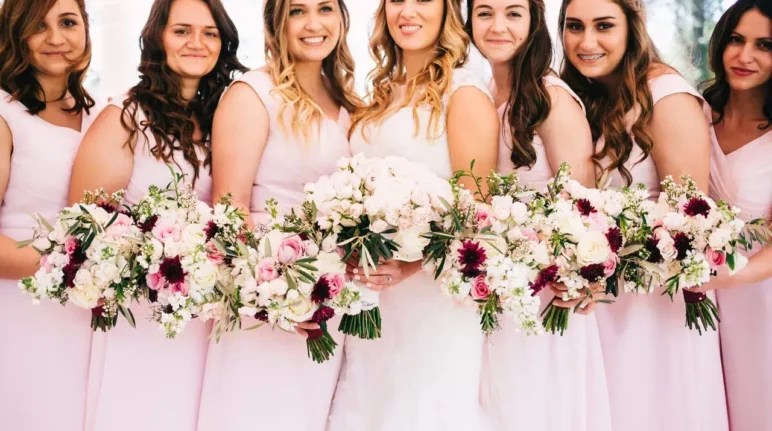 A bride and her bridesmaids, all dressed in soft pink dresses, holding lush bouquets designed by a florist that services weddings, featuring a mix of white and deep pink flowers.