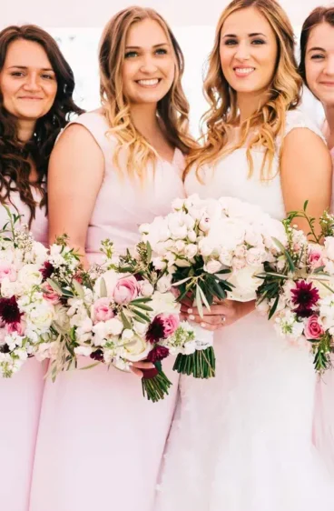 A bride and her bridesmaids, all dressed in soft pink dresses, holding lush bouquets designed by a florist that services weddings, featuring a mix of white and deep pink flowers.