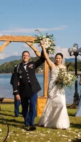 wooden arch by a lake, with the bride holding a bouquet, surrounded by guests and bridesmaids in blue dresses.