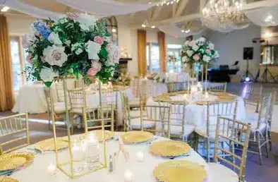 Elegant table setting with tall floral wedding centerpiece featuring roses and greenery, surrounded by gold plates and candles.